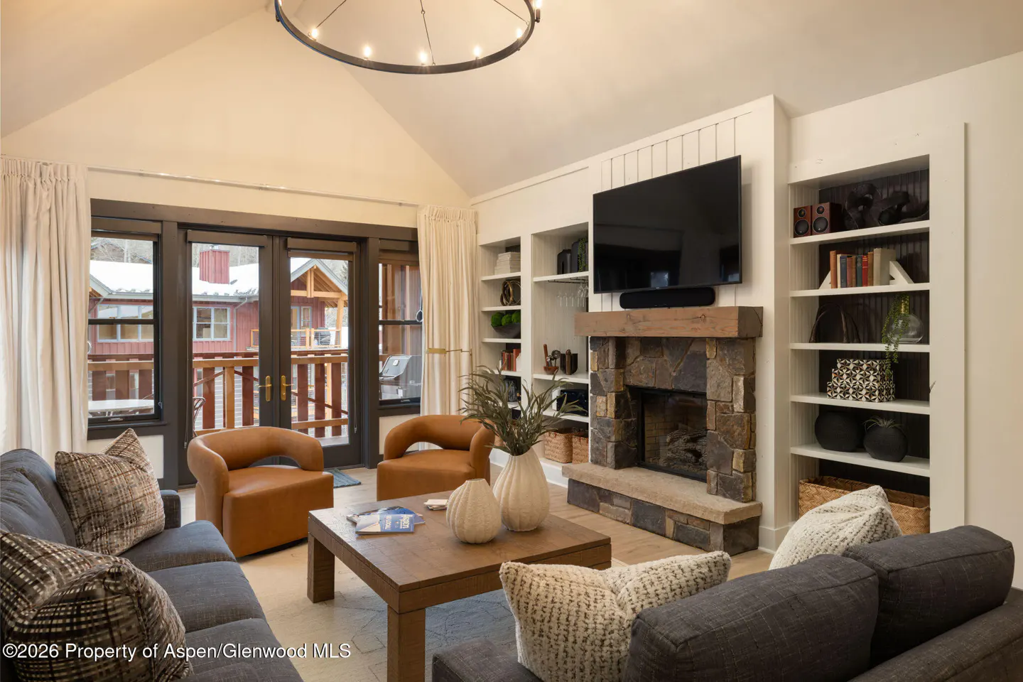 Living room with gray sofas, brown leather chairs, stone fireplace, and built-in bookshelves. Outside, a snowy scene is visible.
