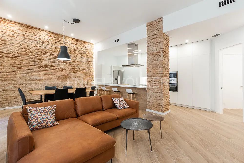 Open-concept living space with exposed brick walls, a brown leather sectional sofa, and a modern kitchen with white cabinets.