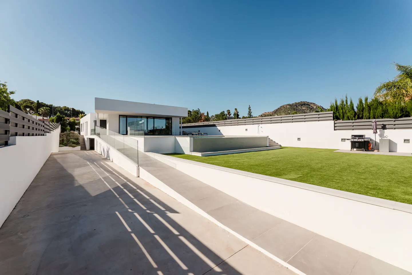 Modern white house with a concrete driveway, glass-railed ramp, green lawn, and a grill against a white perimeter wall.