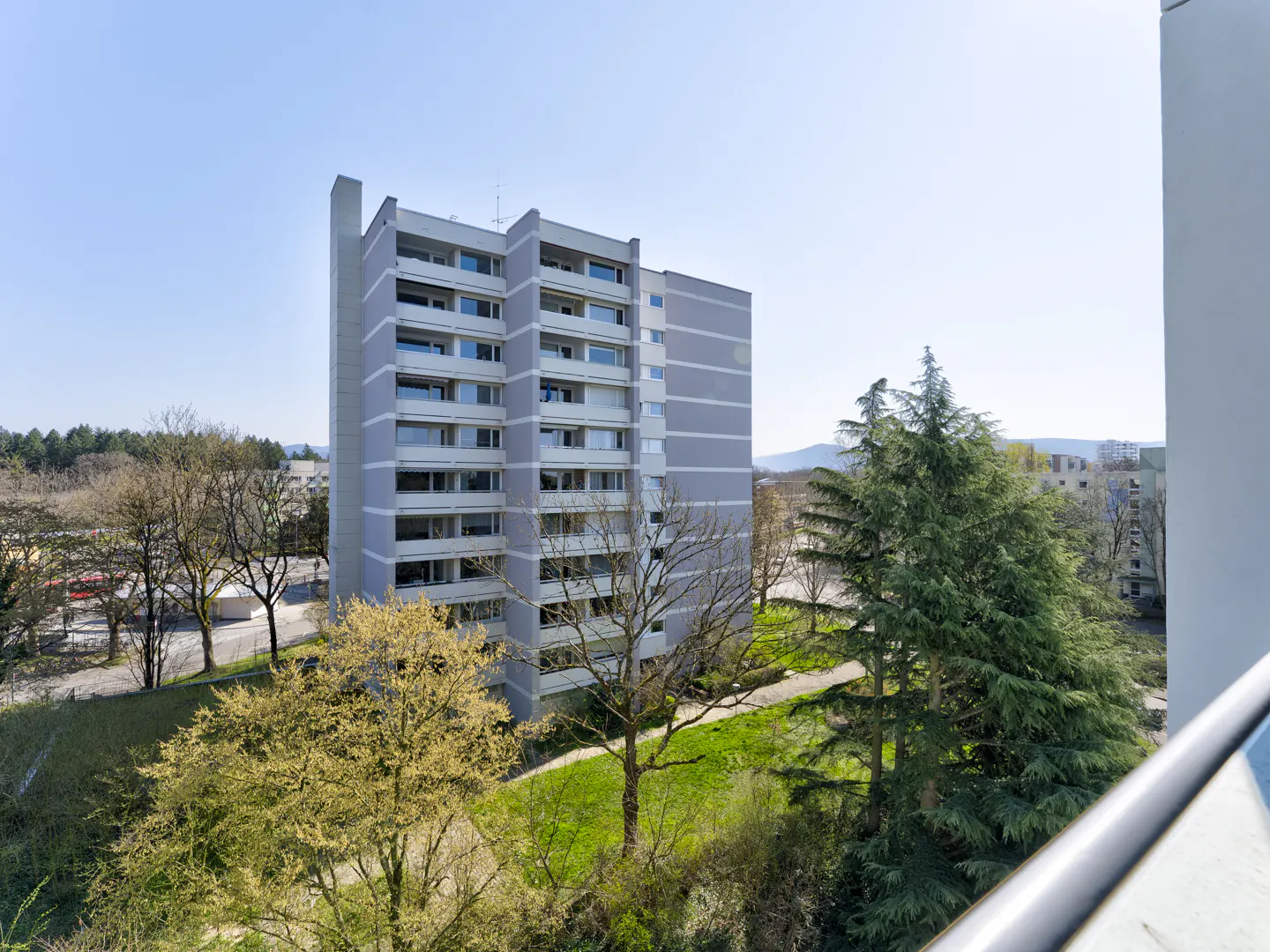 View of a tall, gray apartment building with balconies, surrounded by green trees and grass on a sunny day.