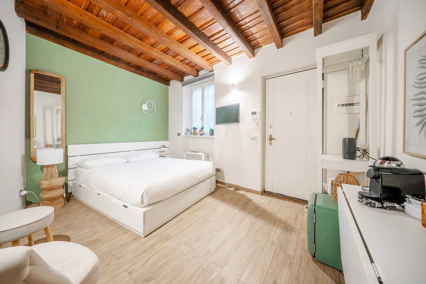 Bedroom with white bed, sage green accent wall, exposed wood ceiling beams, and light wood flooring.