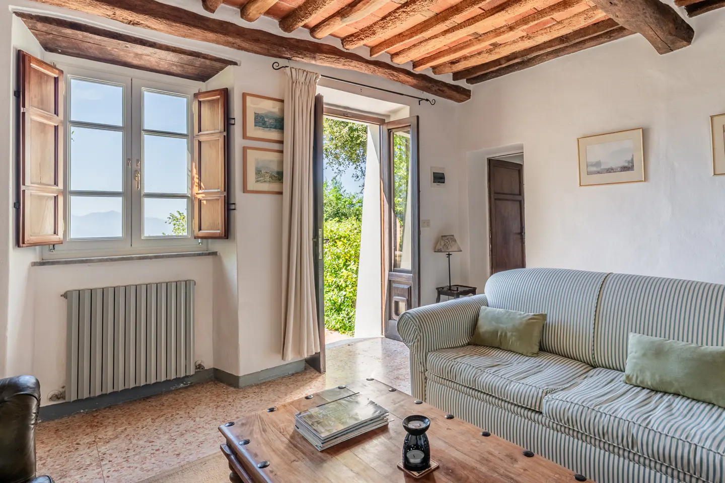 Living room with a striped sofa, wooden table, and open doors leading to a lush garden. The ceiling has exposed wooden beams.