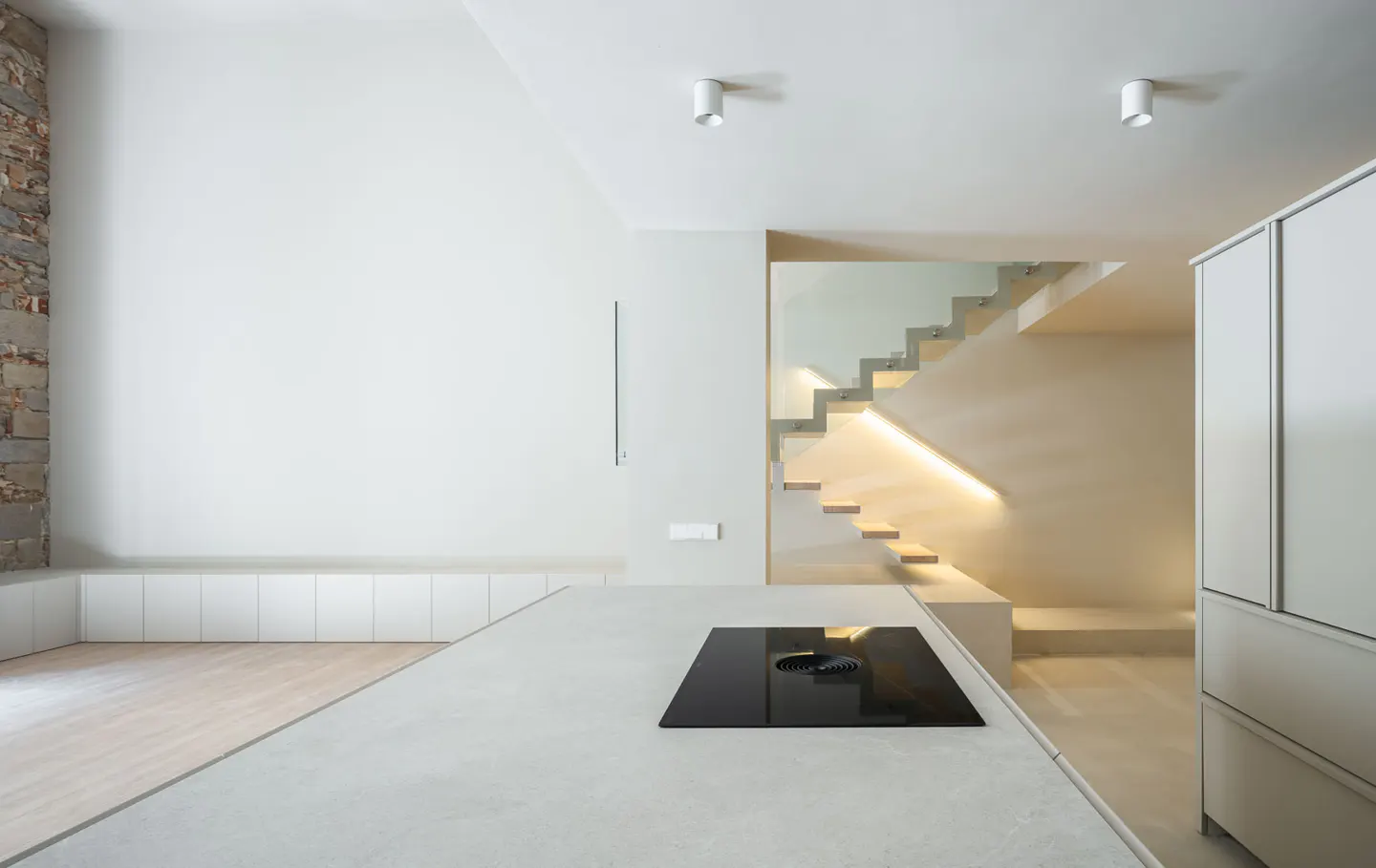 Minimalist kitchen with white walls, cabinets, and stairs with LED lighting. A black stovetop is on the island.