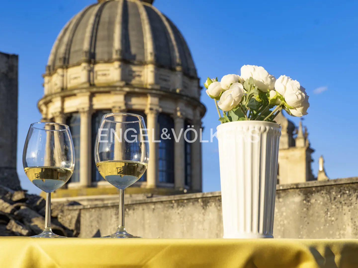 Two glasses of white wine and a vase of white flowers on a table overlooking a historic domed building under a blue sky.