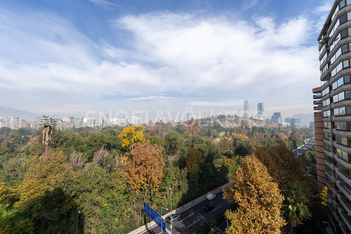 View from a high-rise apartment overlooking a park with autumn foliage, a highway, and the city skyline under a cloudy sky.