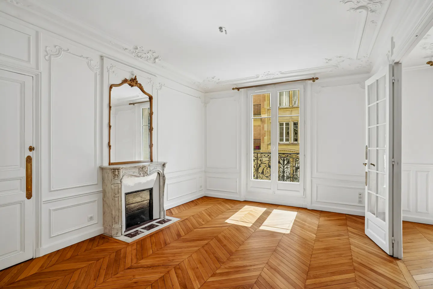 Bright room with white walls, ornate molding, a marble fireplace with a mirror, and herringbone hardwood floors.