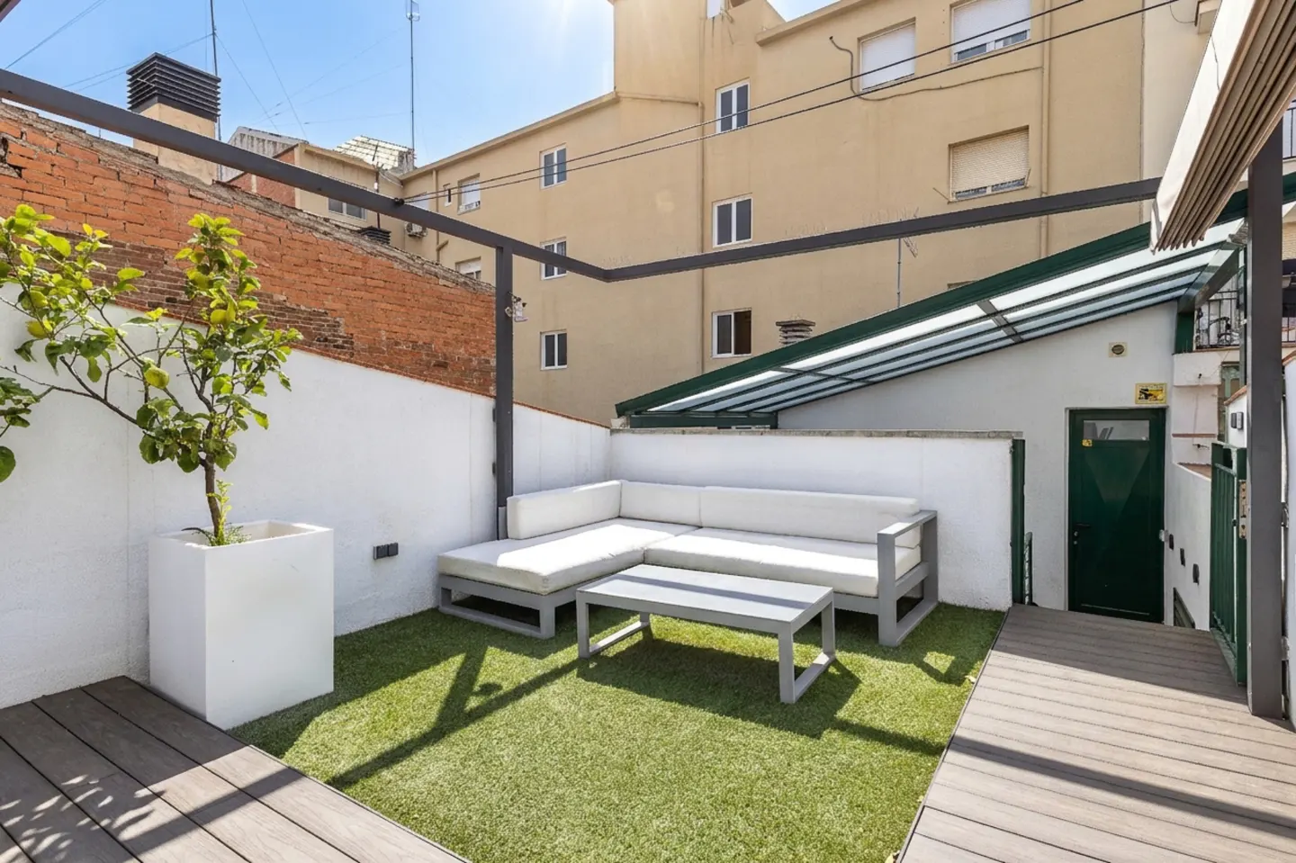 Outdoor patio with white L-shaped sofa, gray table, and green artificial turf. A potted tree stands near a white wall, with buildings in the background.