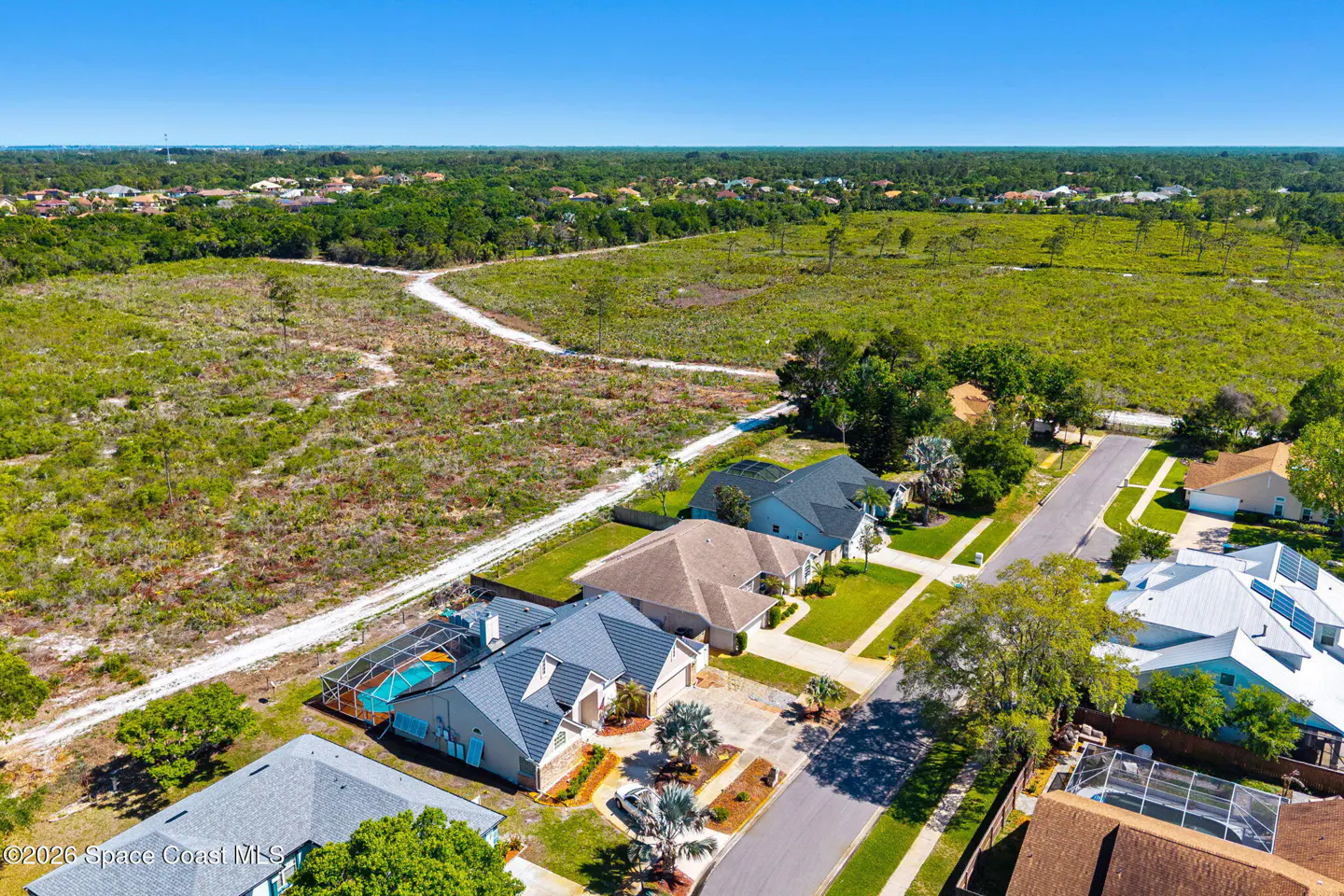 Aerial view of a residential neighborhood with houses, green lawns, and a dirt road leading to a wooded area under a blue sky.