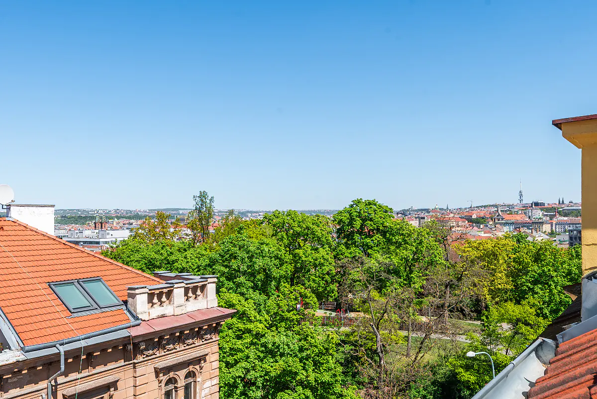 View of Prague from a building with a red tile roof. Green trees fill the midground, and the city skyline is visible in the distance under a clear blue sky.