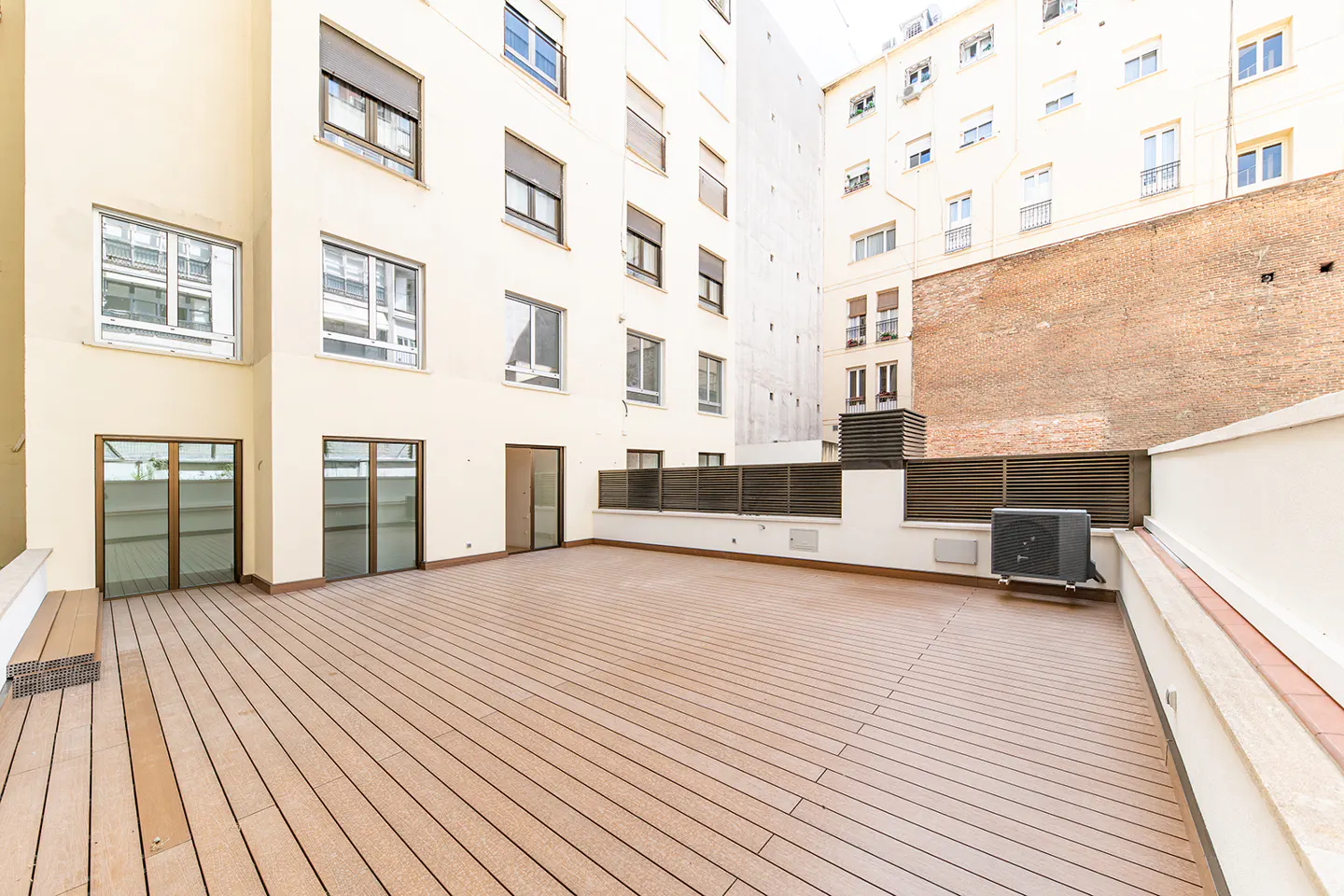 Spacious outdoor terrace with light brown wooden decking, surrounded by tall cream-colored apartment buildings.