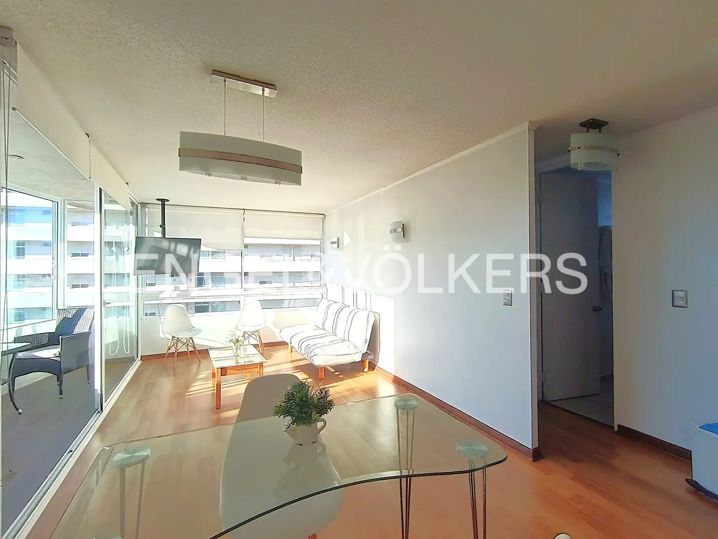 Bright living room with wood floors, glass table, white sofa, and large windows with city views.