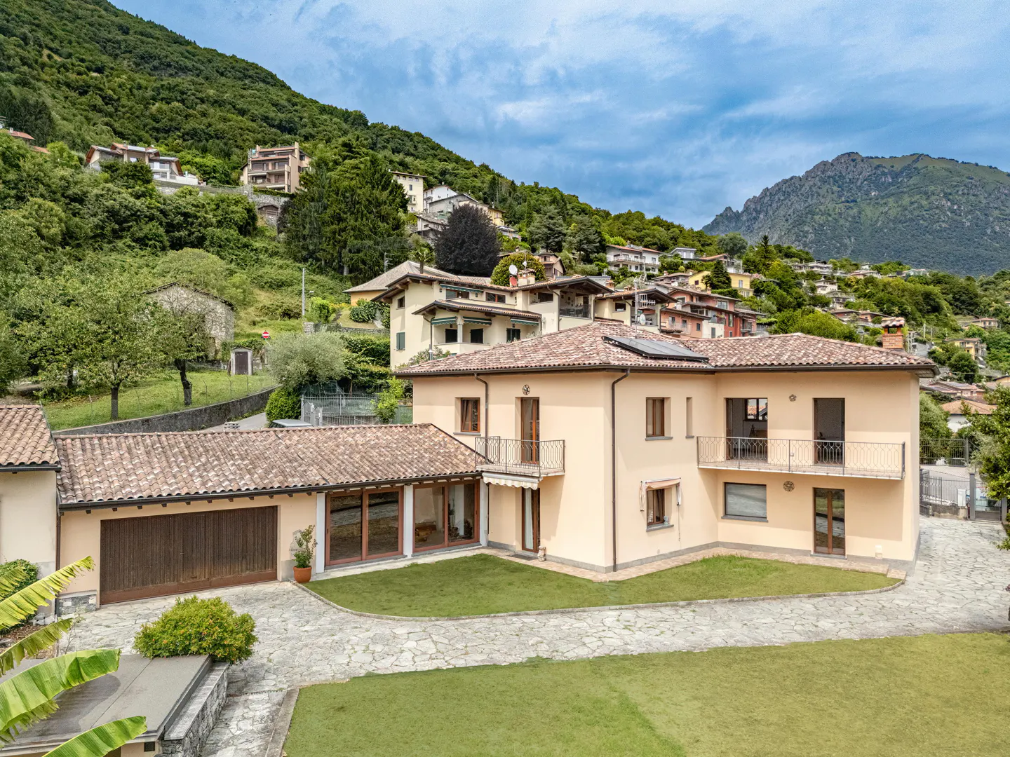 A tan two-story house with a brown tile roof sits on a green lawn with a stone driveway. Balconies adorn the second floor. A mountain covered in trees rises in the background.