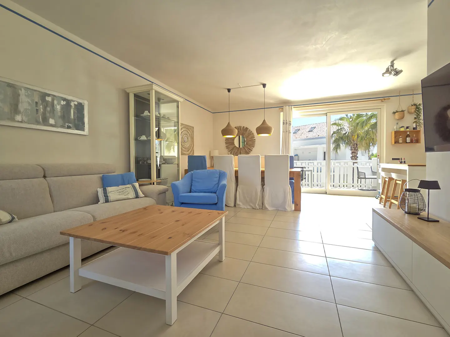 Bright living room with beige sofa, blue armchair, wood coffee table, dining table with white chair covers, and balcony view.