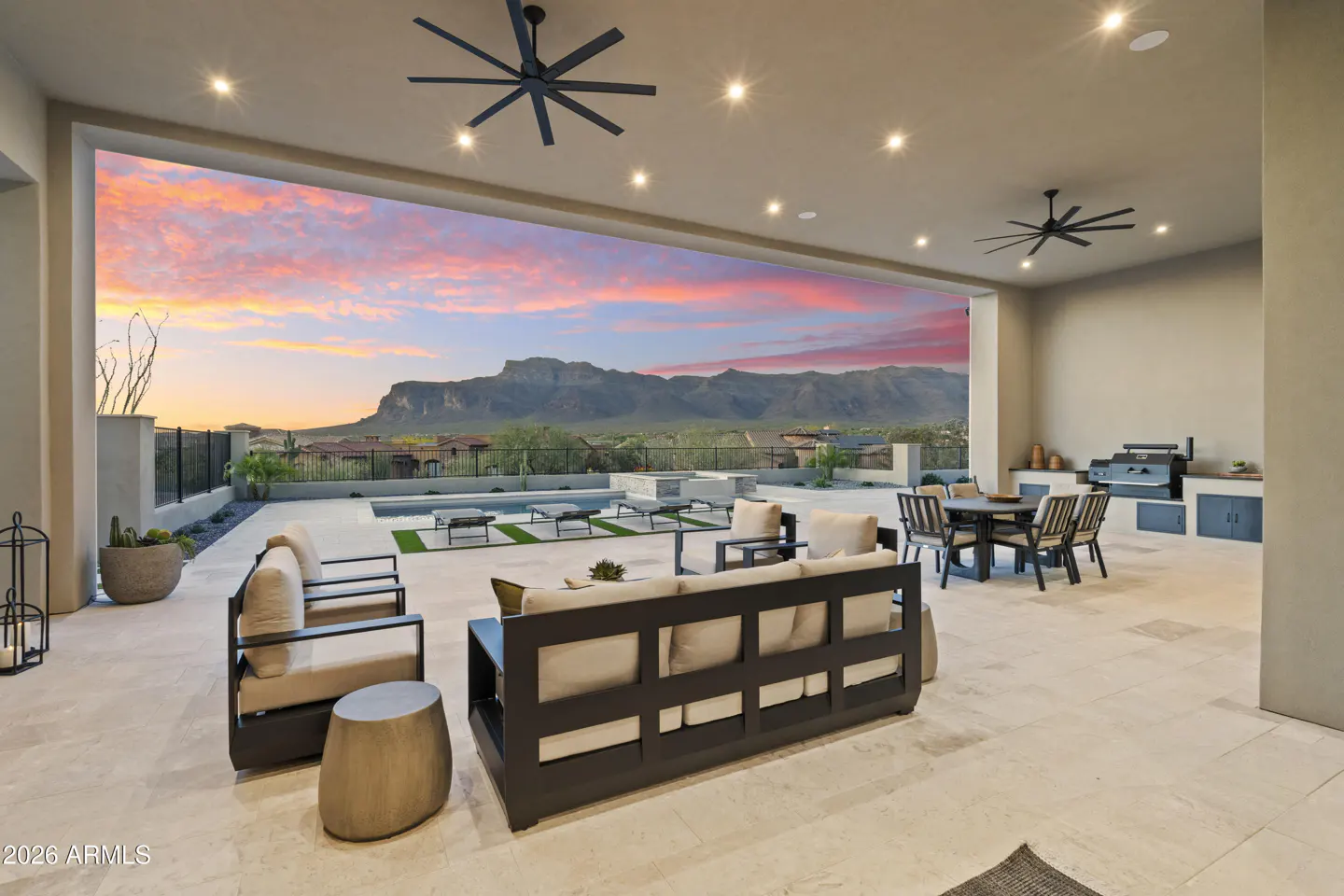 Covered patio with outdoor seating, dining area, and grill overlooking a pool and mountain range at sunset.