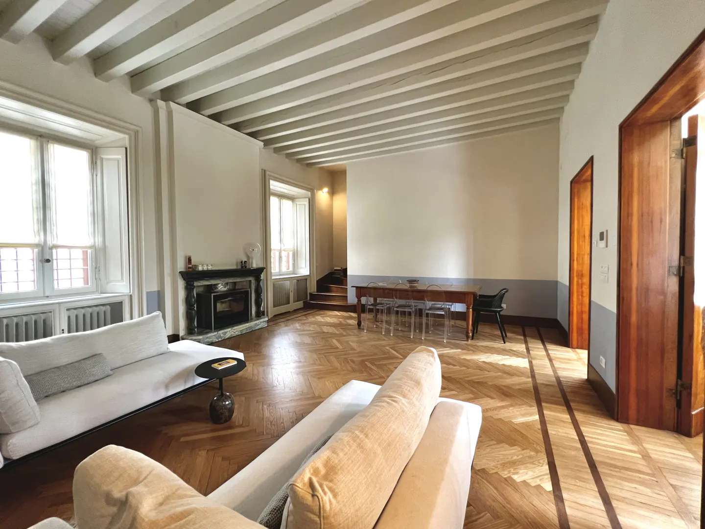 Bright living room with white sofas, herringbone wood floors, and a fireplace. A dining table sits in the background.