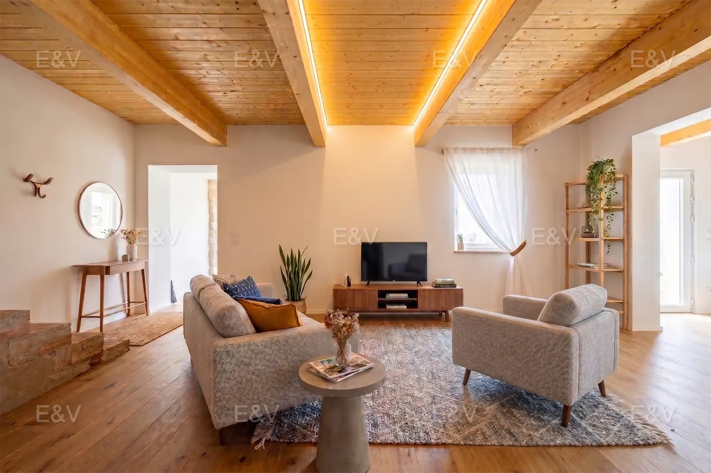 Living room with wood floors, beamed ceiling, and light gray sofa and armchair. A TV sits on a wood console.