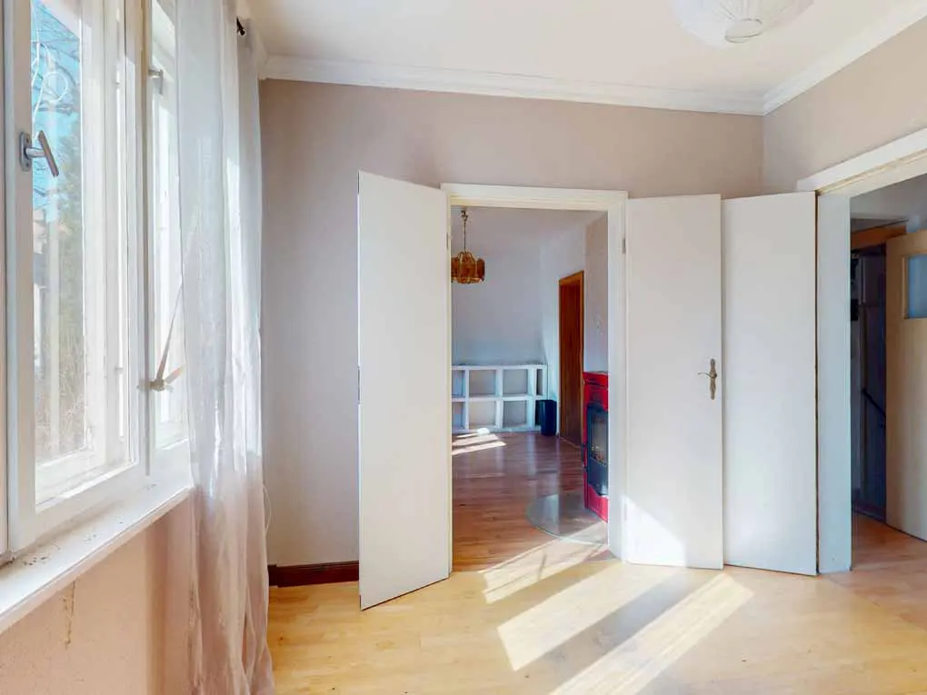 A bright room with hardwood floors, white walls, and open double doors leading to another room. A window with sheer curtains is on the left.
