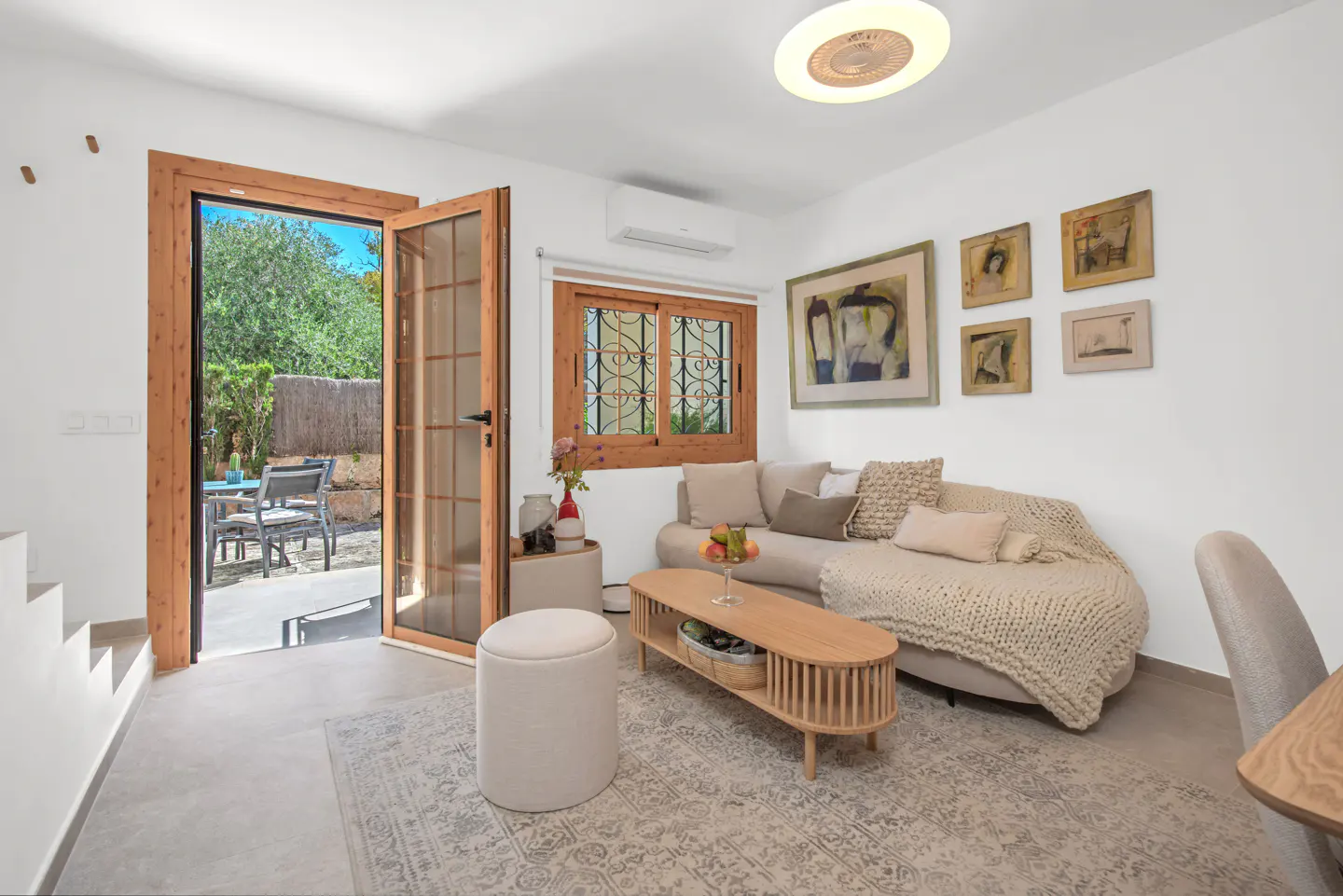 Bright living room with white walls, a beige sofa, and a wooden coffee table. Open doors lead to a patio with outdoor seating.
