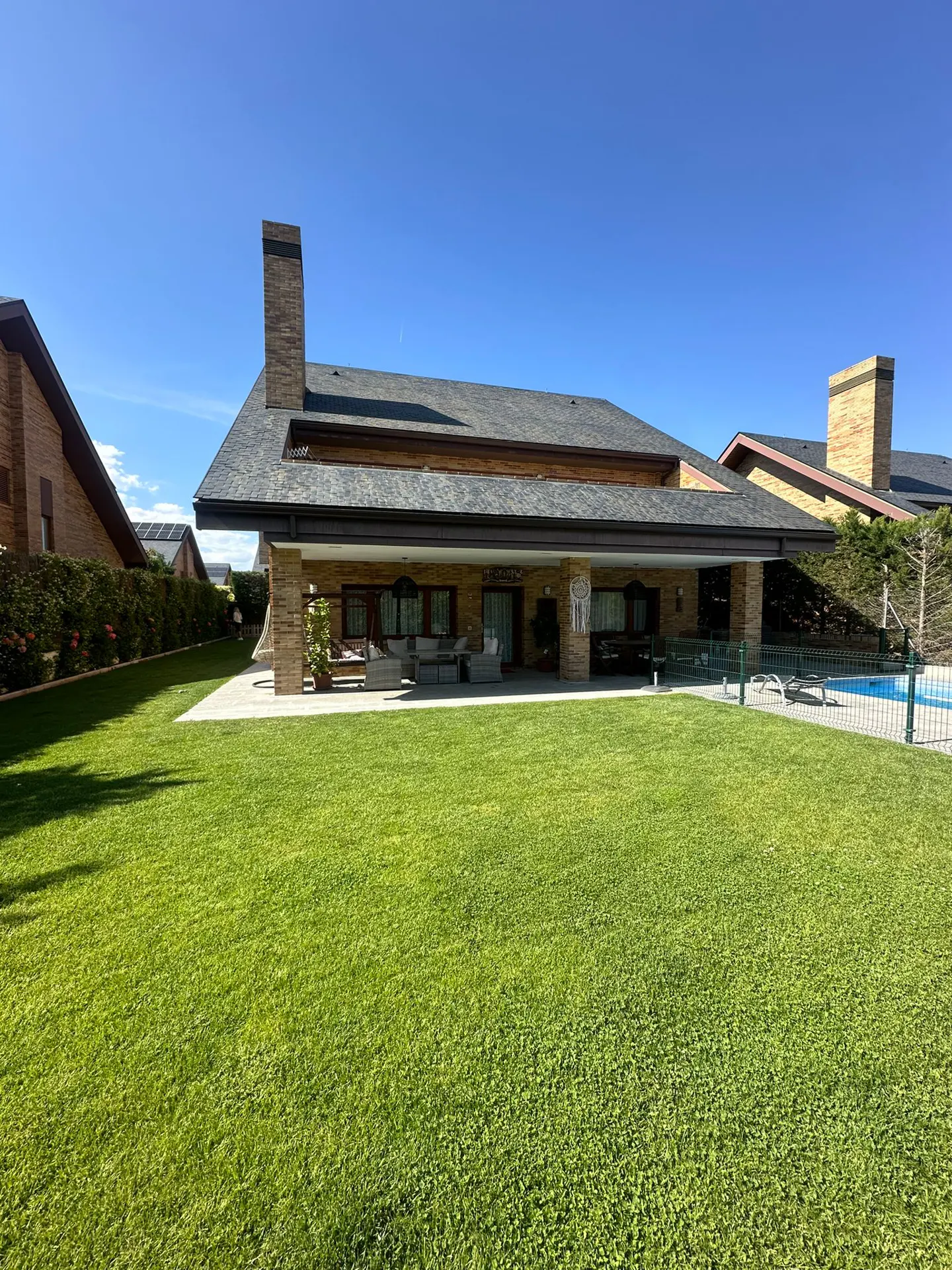 Backyard view of a tan brick house with a gray roof, chimney, patio furniture, green lawn, and a fenced-in pool under a blue sky.