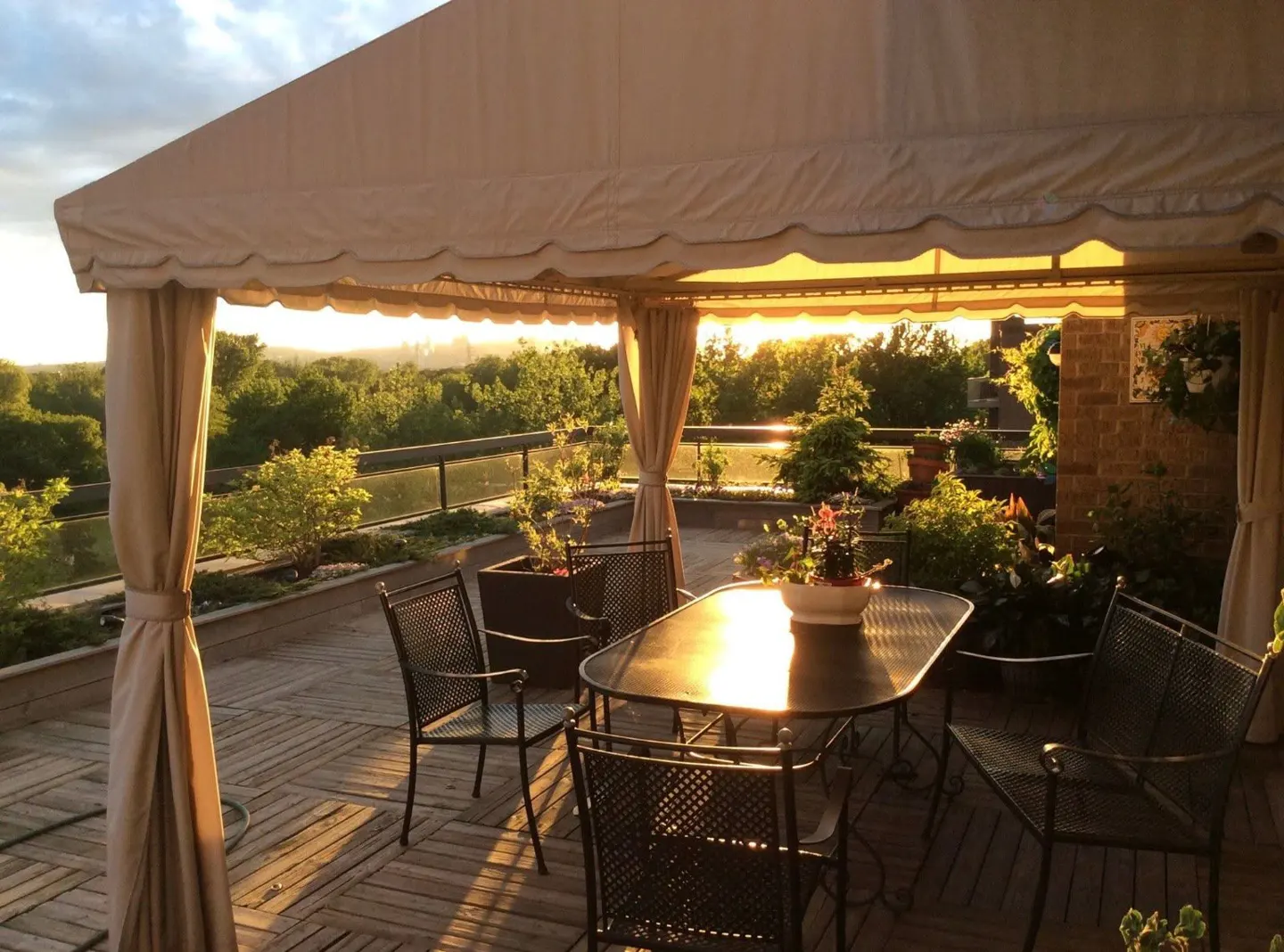 Outdoor patio with a table and chairs under a beige canopy, overlooking a green landscape at sunset.