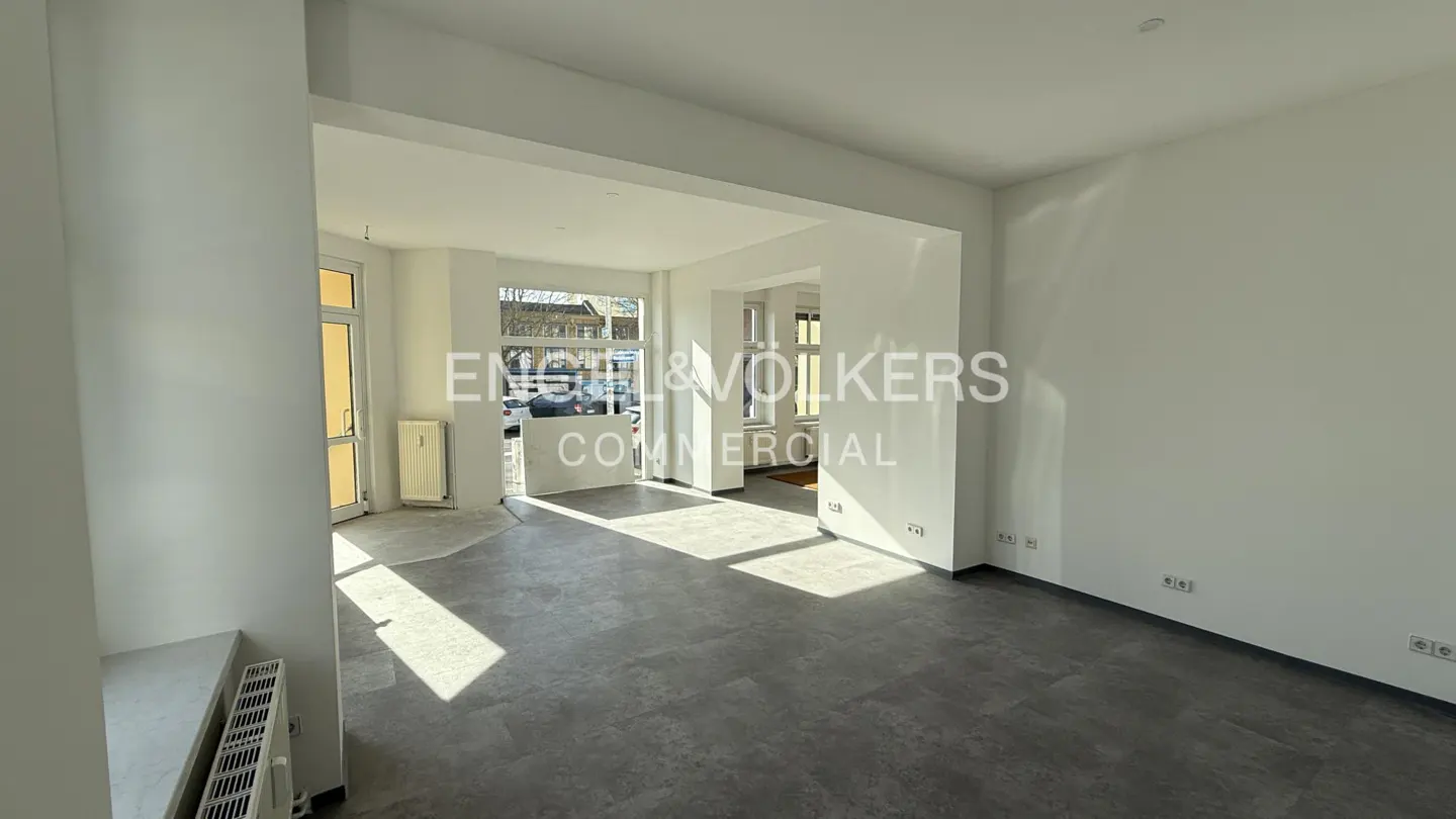 Bright, empty commercial space with gray tile floor, white walls, and open doorway to the street. Engel & Volkers logo is superimposed.