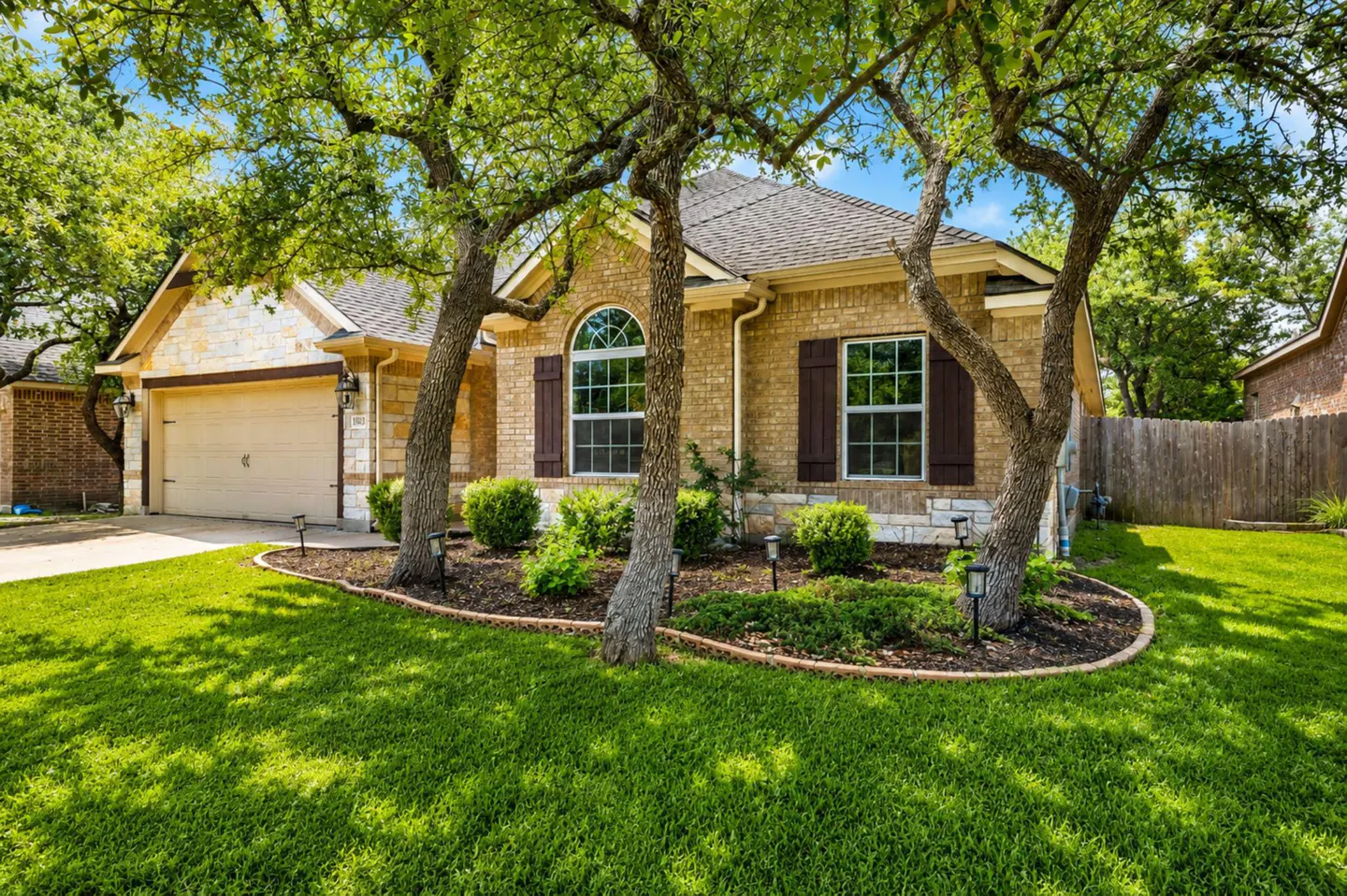 A tan brick house with brown shutters is framed by trees and green lawn.