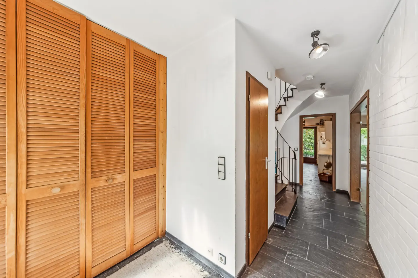 Hallway with wood closet, slate floor, and white brick wall. A staircase and doorways lead to other rooms.
