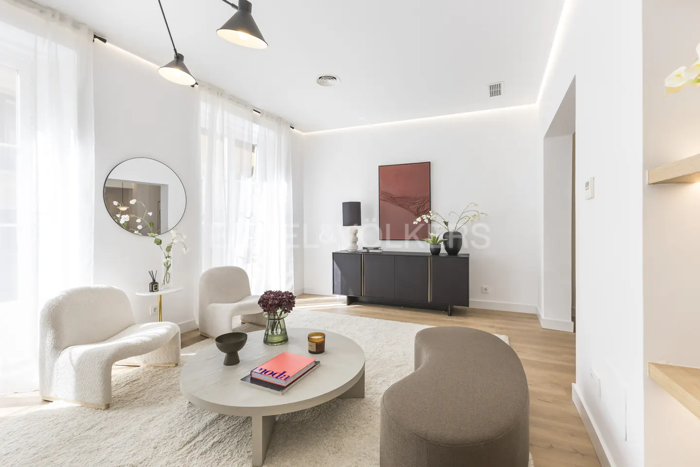Bright living room with white walls, light wood floors, and a cream rug. Two white chairs face a round coffee table. A black cabinet sits against the wall.
