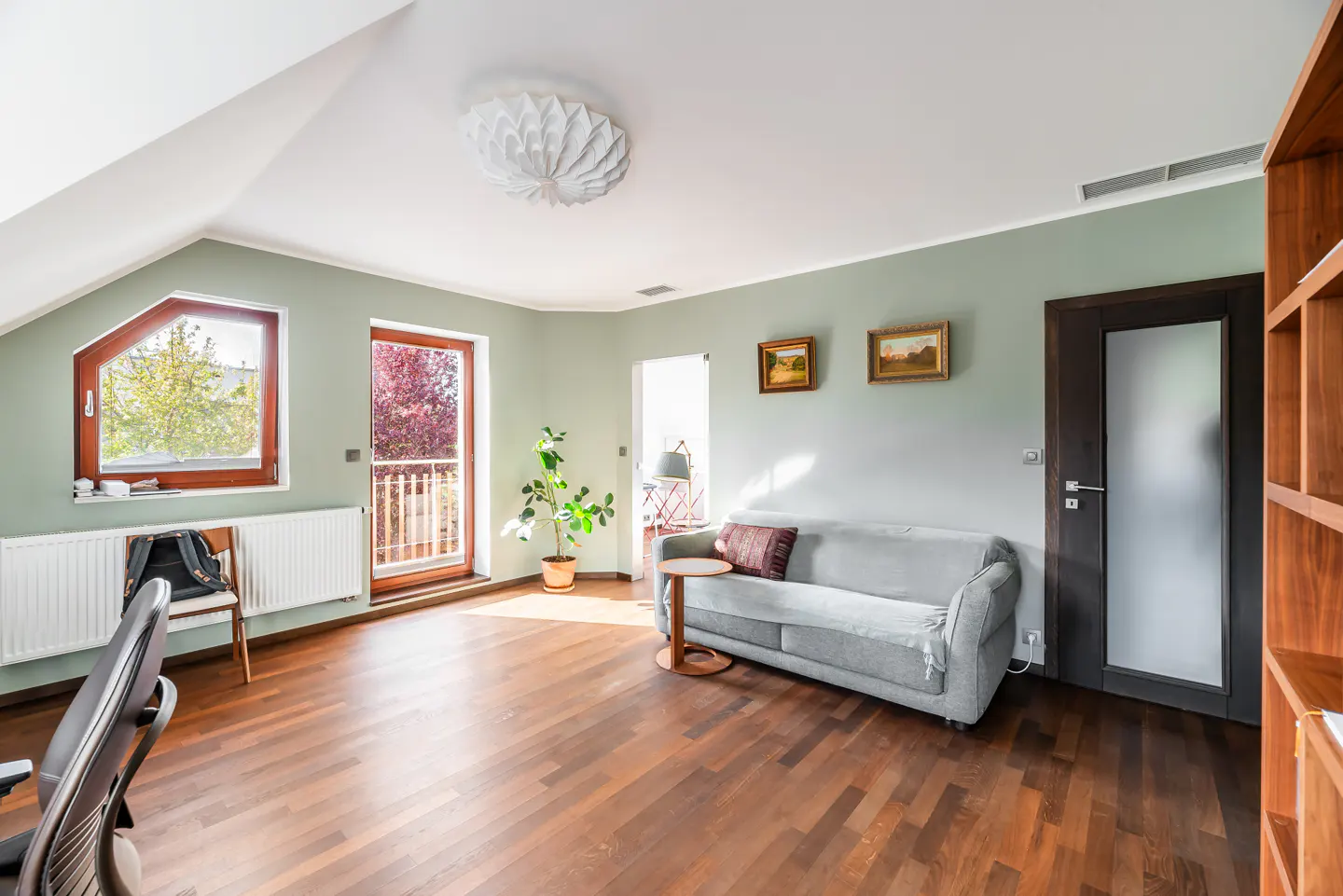 Attic room with sage green walls, wood floors, and a gray sofa. A window and door lead to the outside. Two paintings hang above the sofa.