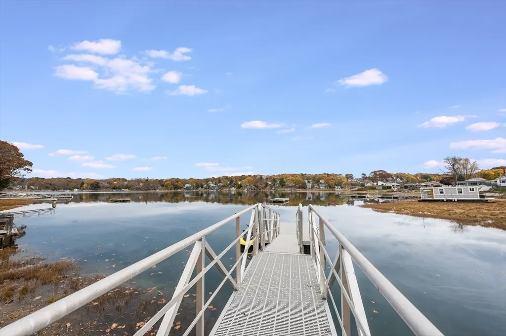 A metal dock extends into a calm lake under a blue sky, with autumn trees and houses visible along the distant shoreline.