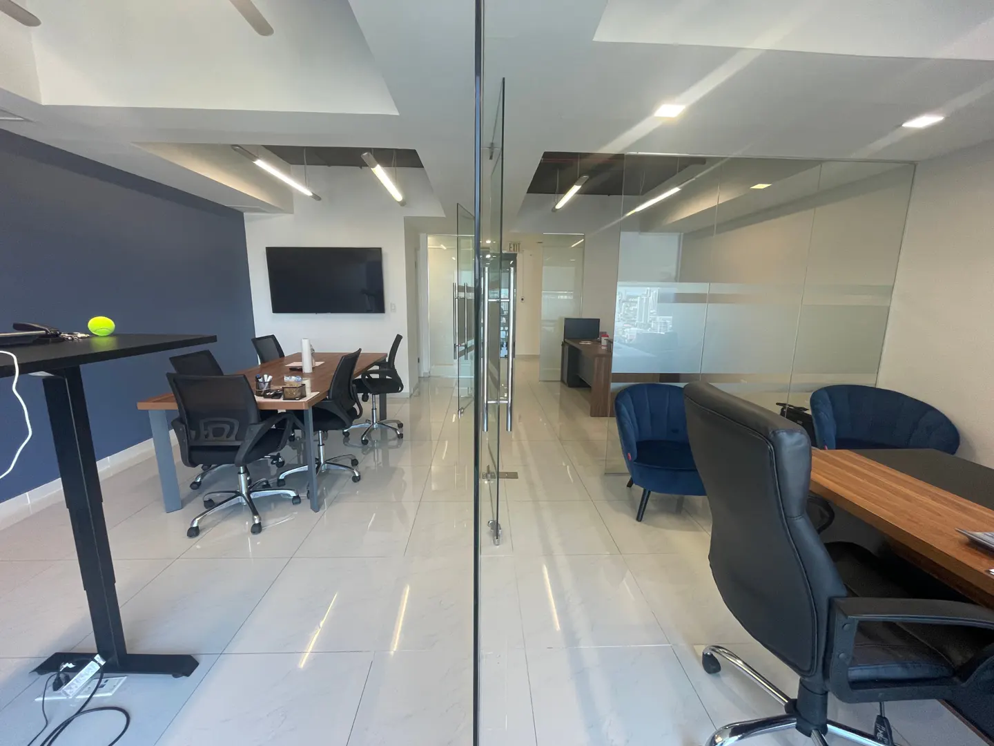 A modern office space with glass walls, a conference table with black chairs, and blue accent chairs. A black TV is mounted on a blue wall.