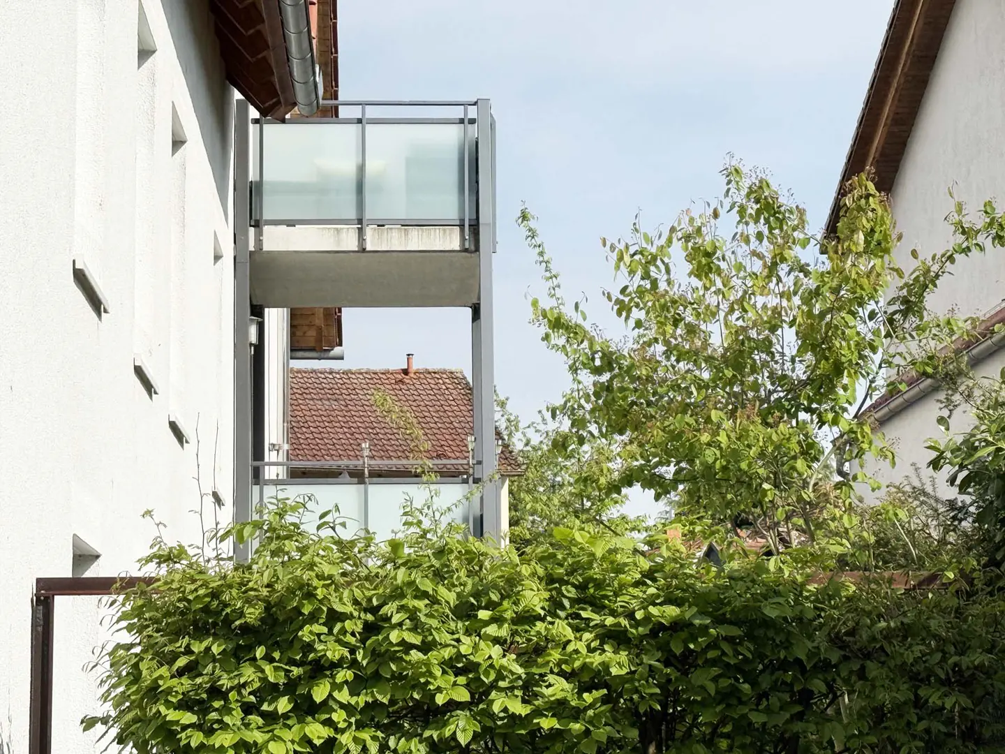 Exterior view of a white building with balconies, green bushes, and a red-tiled roof in the background.