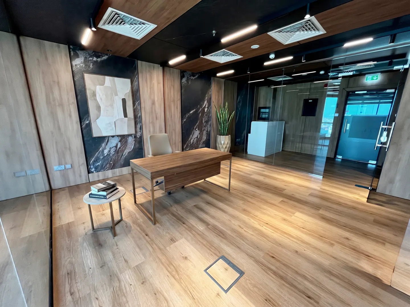Office interior with wood floors, walls, and desk. A glass-walled room is visible in the background. A small table with books is in the foreground.