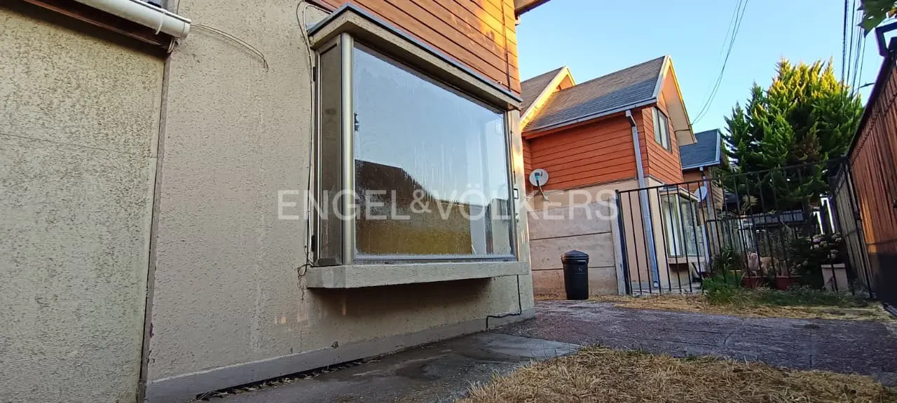 Exterior view of a beige stucco house with a large bay window and the Engel & Volkers logo. A neighboring orange house is visible.