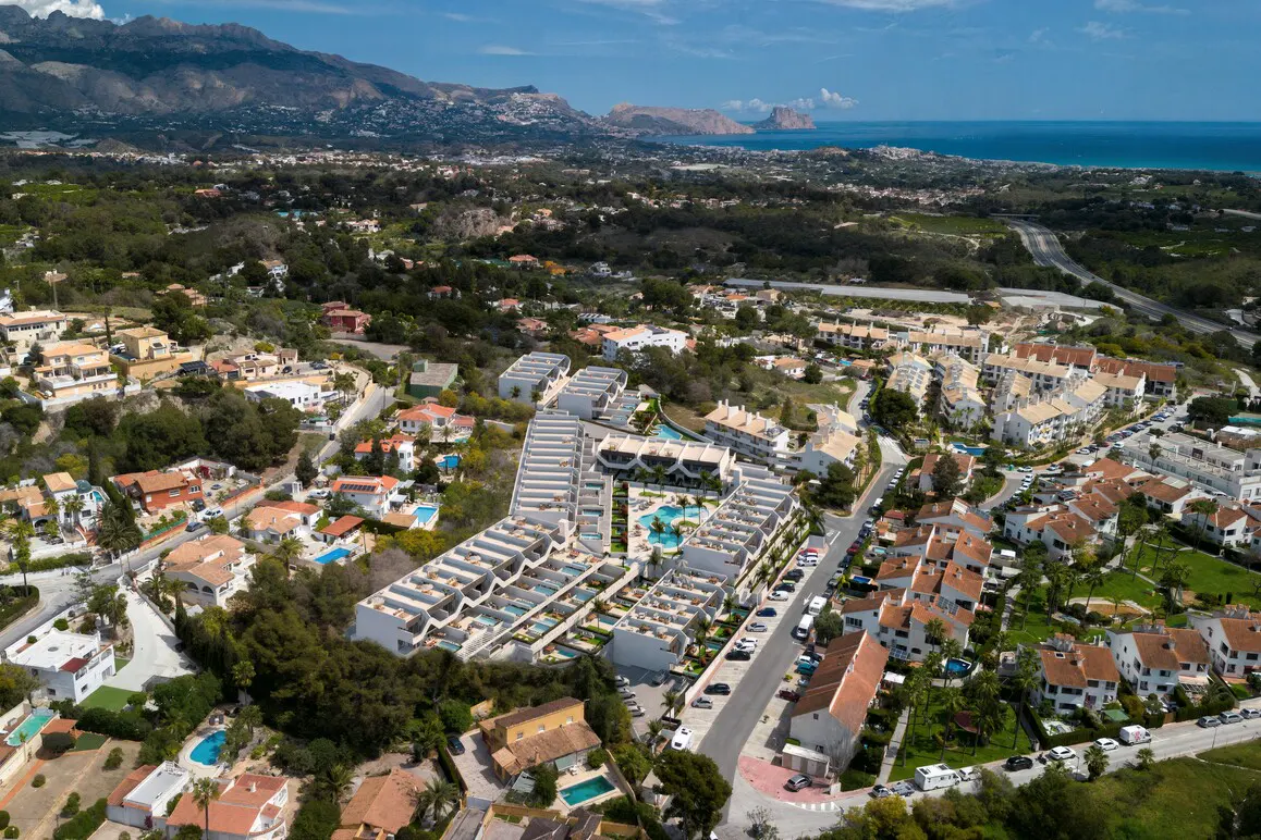 Aerial view of a white apartment complex with pools, surrounded by houses, trees, and mountains under a blue sky.