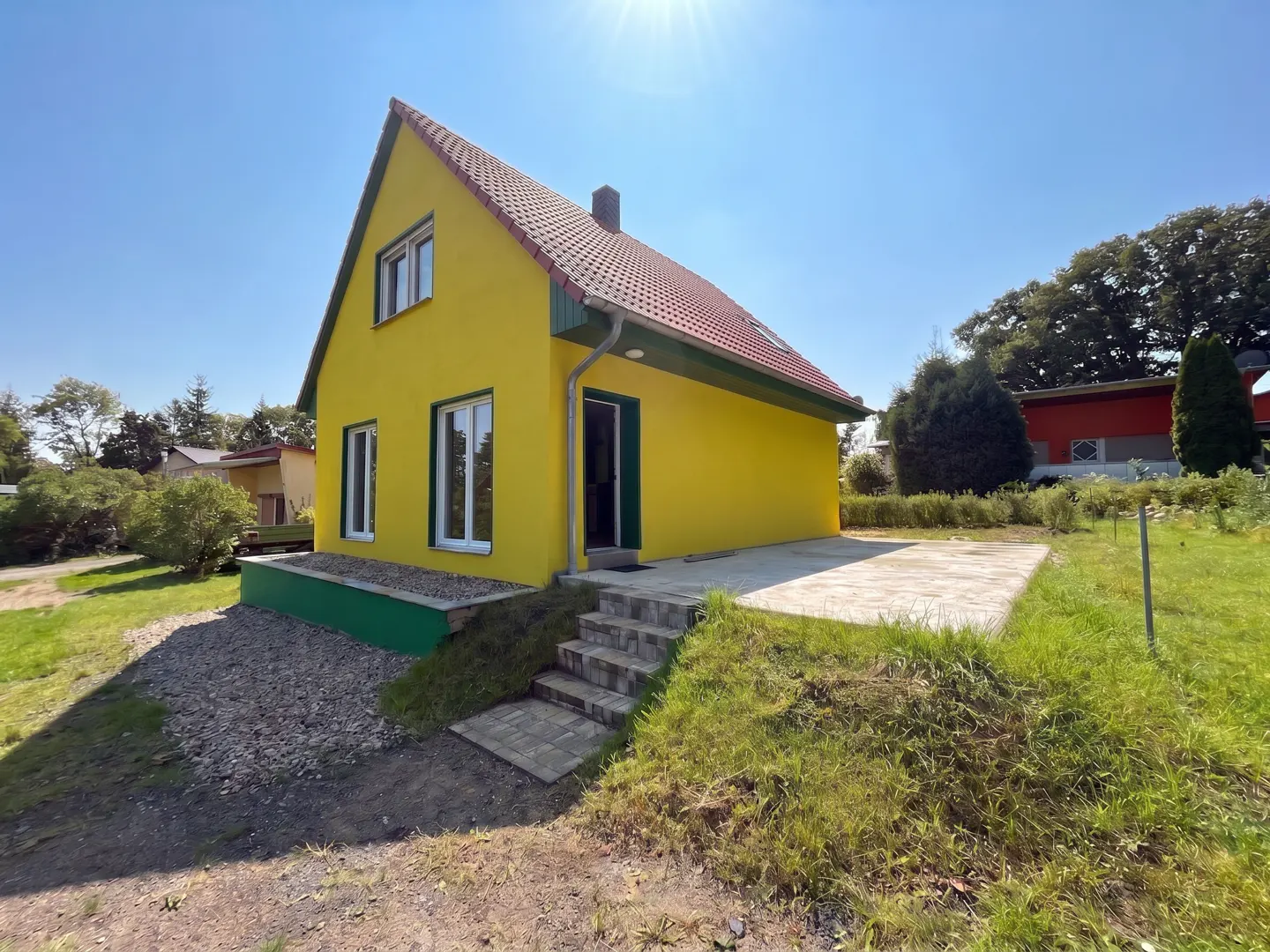 A yellow, two-story house with a red roof, green trim, and a stone patio on a sunny day.