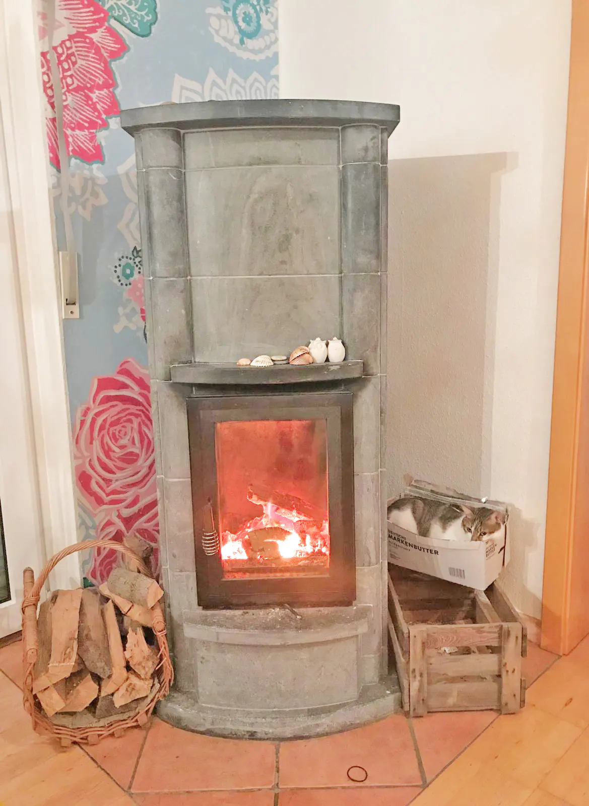 Cozy living room with a grey stone fireplace, burning logs, a basket of wood, and a cat sleeping in a box nearby.