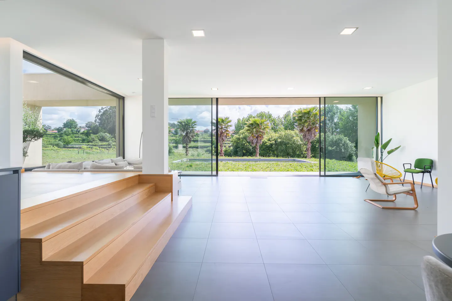 Bright, modern living room with gray tile floors, wooden steps, and large windows overlooking a green lawn and trees. Chairs are visible.
