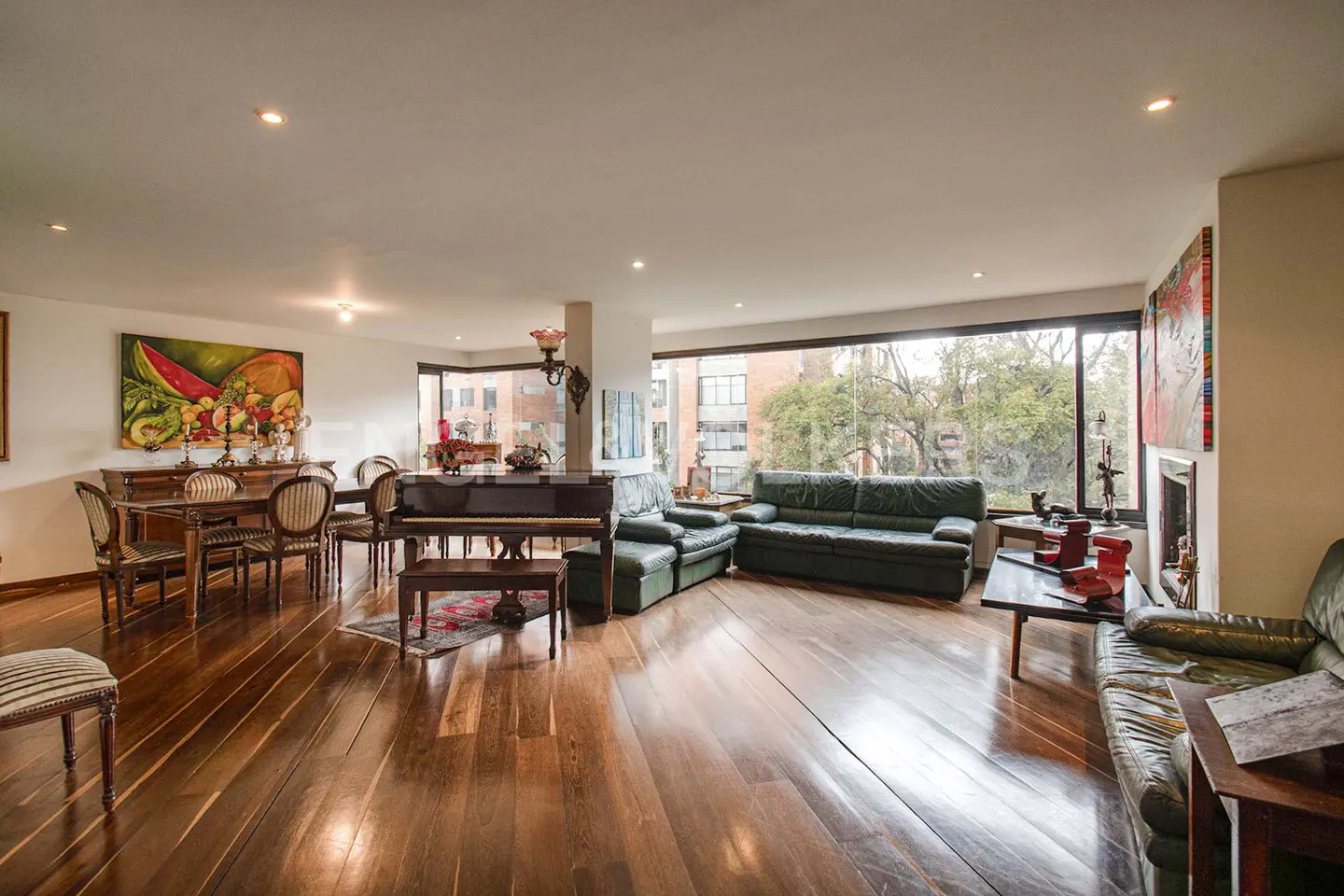 Bright living room with hardwood floors, a dining table, a piano, and green leather sofas. Large windows offer a view of trees.