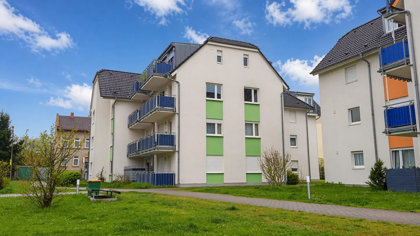 Exterior view of a modern apartment building with blue balconies and green accents, set against a blue sky with scattered clouds.