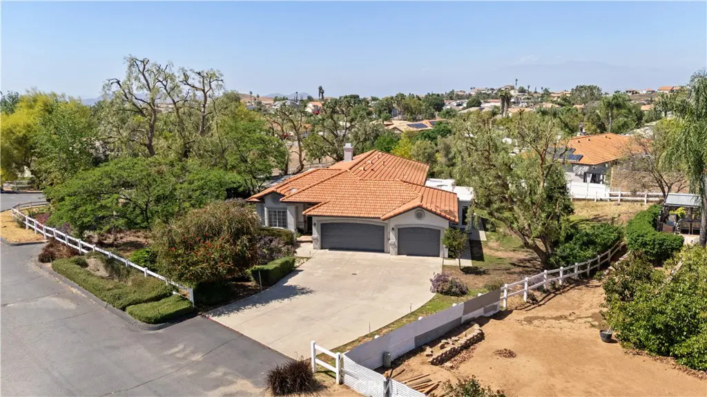Aerial view of a gray house with a terracotta roof, a two-car garage, and a long driveway surrounded by green trees and a white fence.
