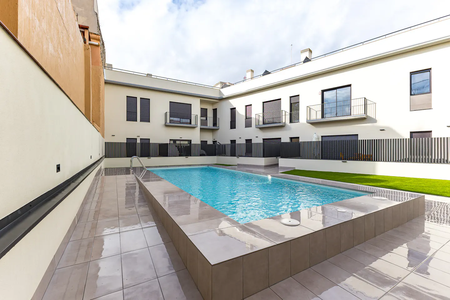 A rectangular swimming pool with turquoise water, surrounded by a tiled patio and white buildings.