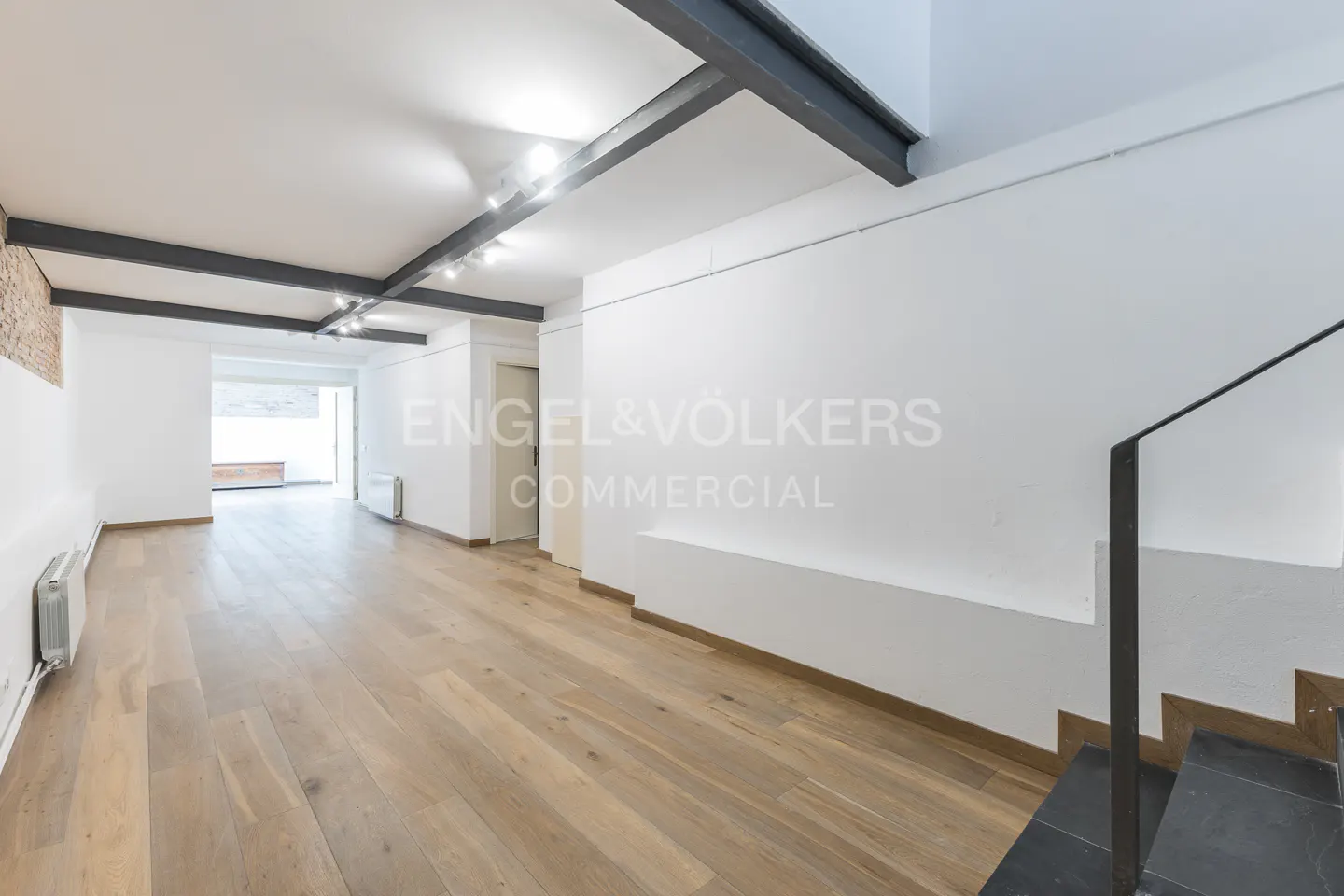 Bright, empty room with hardwood floors, white walls, and black ceiling beams. Stairs with black steps are on the right. "Engel & Völkers" logo on the wall.