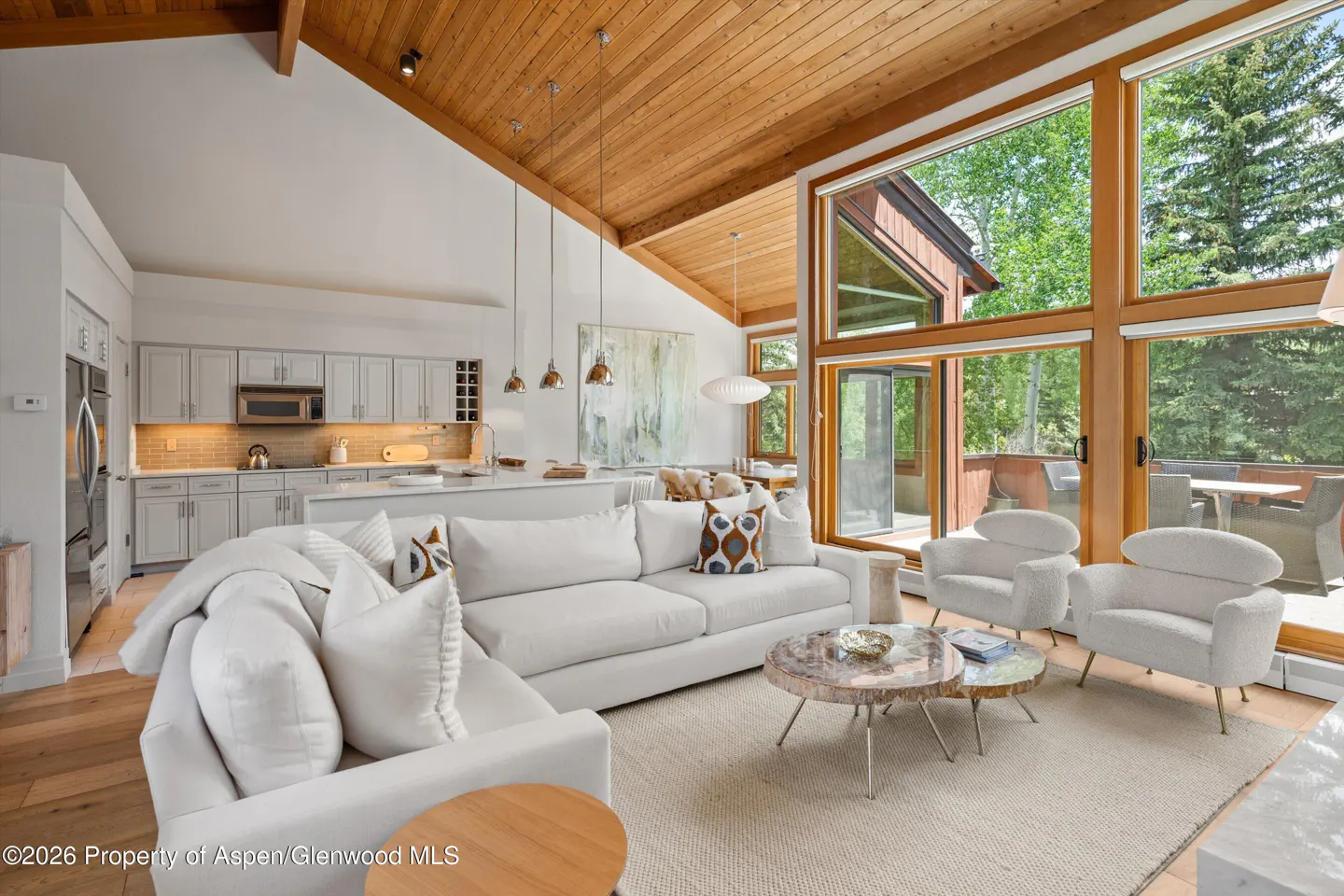 Modern living room with vaulted wood ceiling, white sectional, two armchairs, and floor-to-ceiling windows overlooking trees.