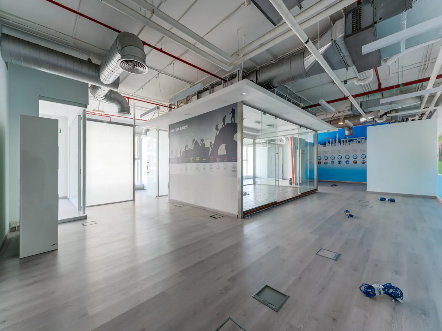 Bright, empty office space with gray wood floors, glass-walled rooms, and exposed ductwork. A motivational poster is on the wall.