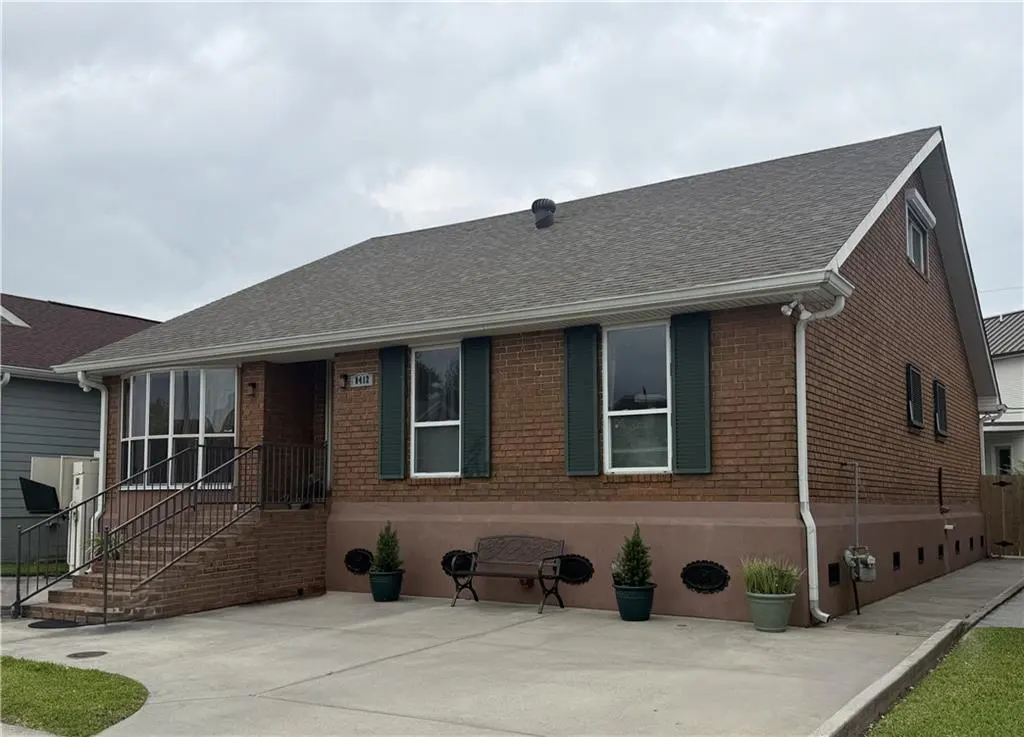 A single-story brick house with green shutters, a brown roof, and a concrete driveway. A bench sits in front of the house.
