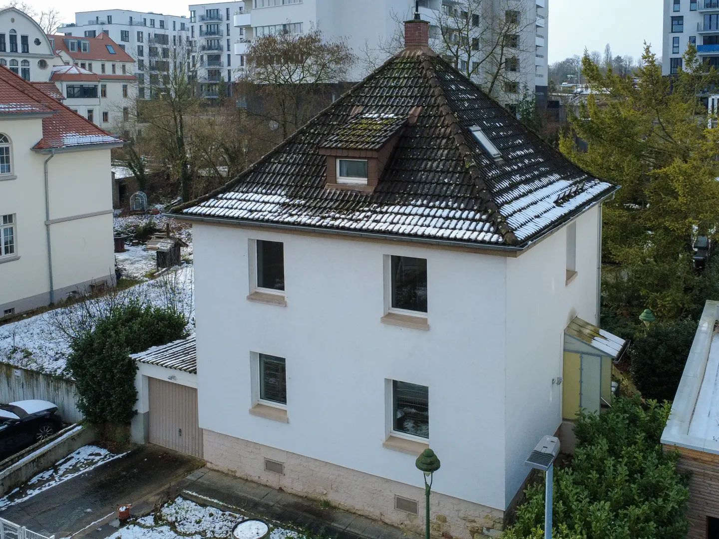 Two-story white house with a brown tiled roof and four windows, surrounded by trees and other buildings.