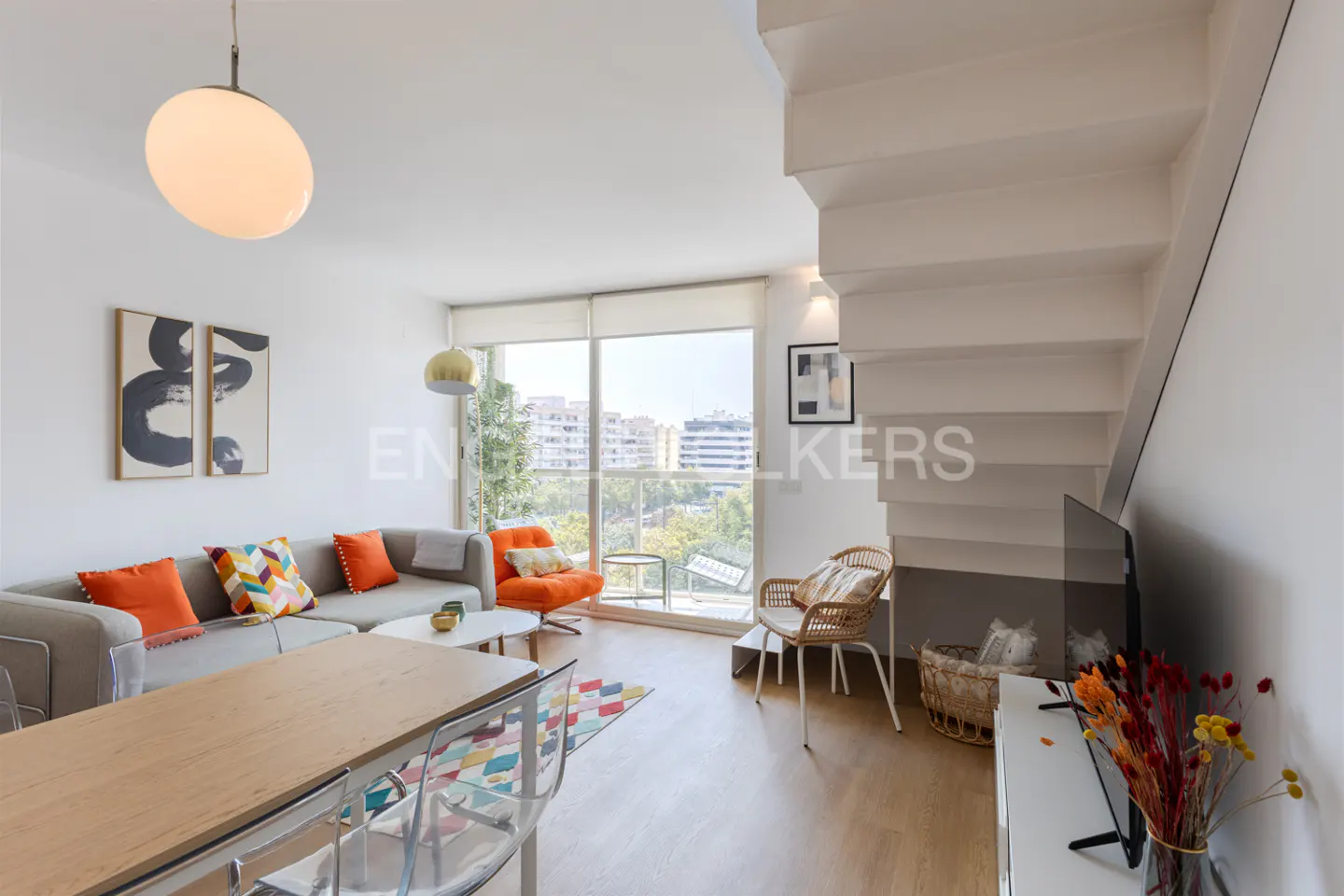 Bright living room with gray sofa, orange chair, wood table, and white stairs. Large window with city view.