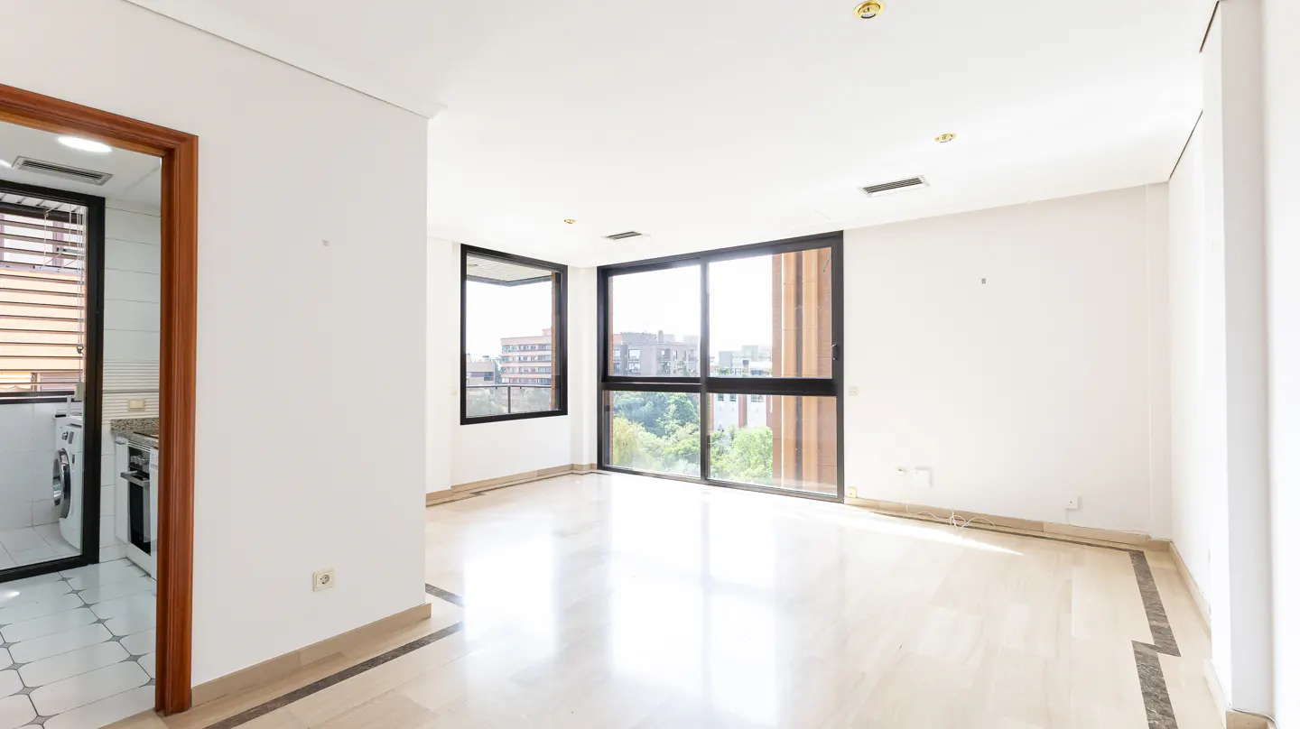 Bright, empty room with marble floors, white walls, and black-framed windows overlooking a city. A doorway reveals a kitchen with white appliances.