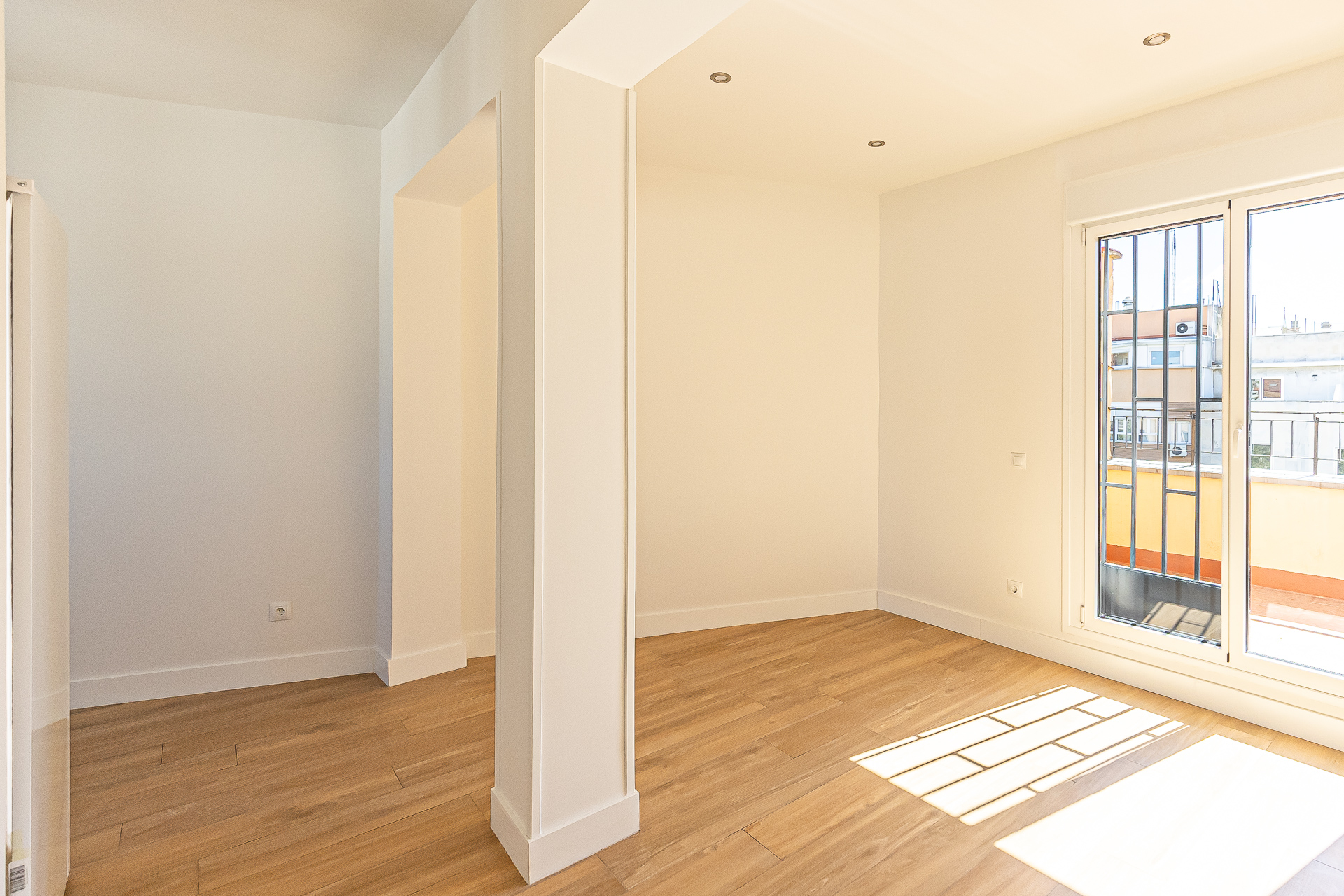 Bright, empty room with light wood floors, white walls, and a large window with bars. Sunlight streams in, creating shadows on the floor.